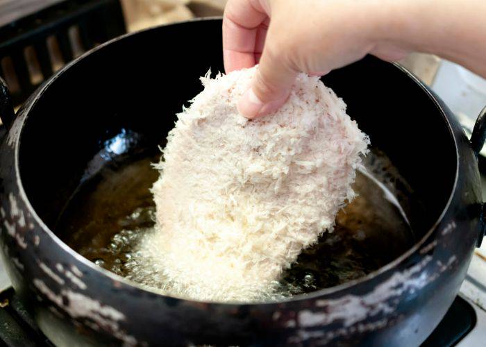 A breadcrumbed tonkatsu pork cutlet being gently placed into hot oil to deep-fry.